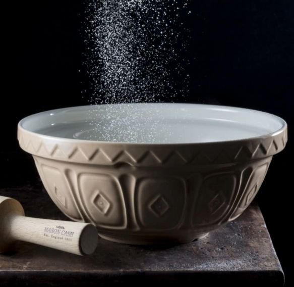 Ceramic mixing bowl with flour being sifted above it, wooden pestle nearby, on rustic dark background.