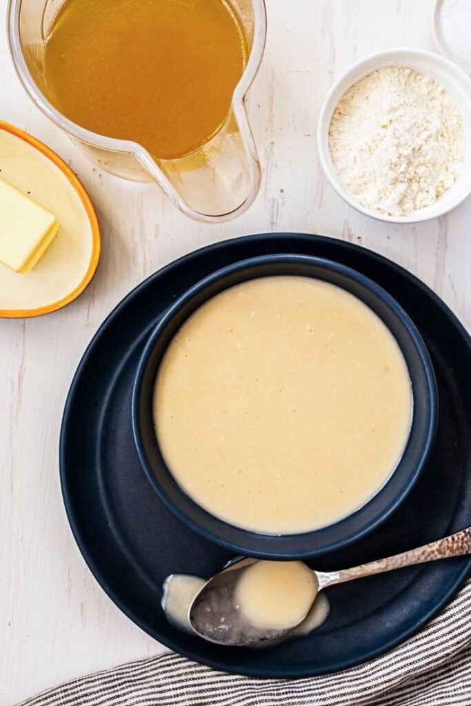 Creamy homemade gravy in a black bowl with ingredients: broth, butter, and flour on a white table.