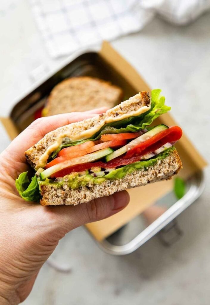 Hand holding a fresh veggie sandwich with lettuce, tomato, cucumber, and avocado on whole grain bread.