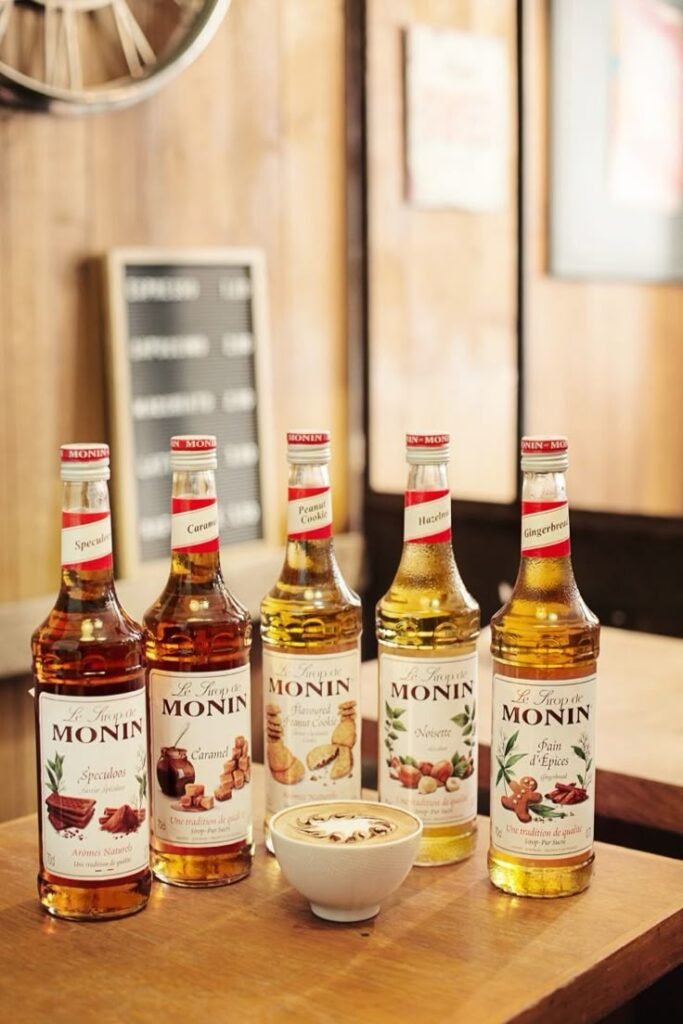 Five Monin syrup bottles and a cappuccino on a wooden table in a cozy café setting.