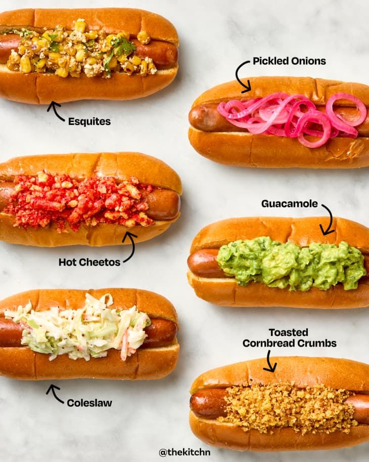 Gourmet hot dogs with toppings: esquites, pickled onions, Hot Cheetos, guacamole, coleslaw, and cornbread crumbs.