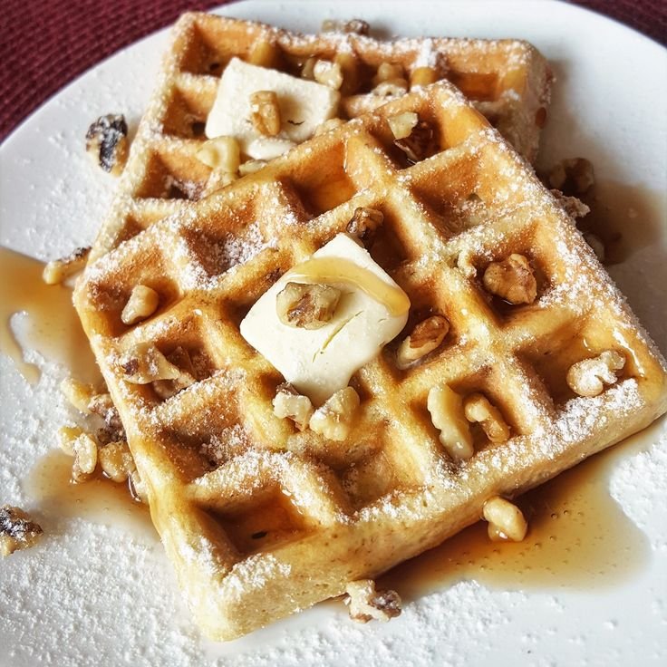 Golden waffles topped with butter, syrup, and walnuts on a plate, dusted with powdered sugar. Delicious breakfast treat.