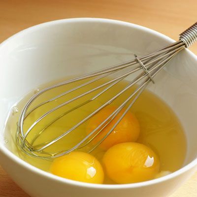 Whisk and three eggs in a white bowl on a wooden surface, ready for mixing in baking or cooking preparation.