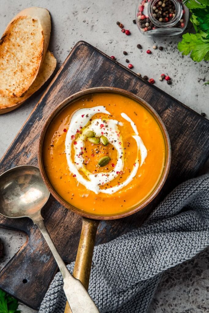 A bowl of creamy pumpkin soup garnished with seeds and cream, served with bread and peppercorns on a rustic board.