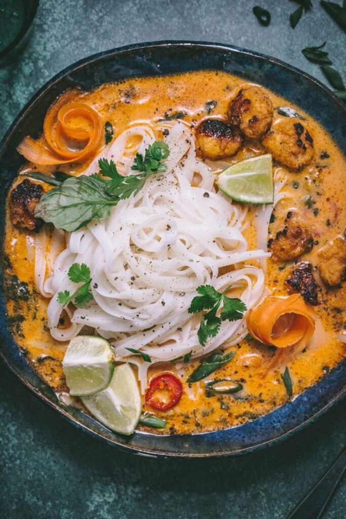 Delicious Thai curry noodle soup with shrimp, lime wedges, and fresh herbs in a flavorful orange broth.