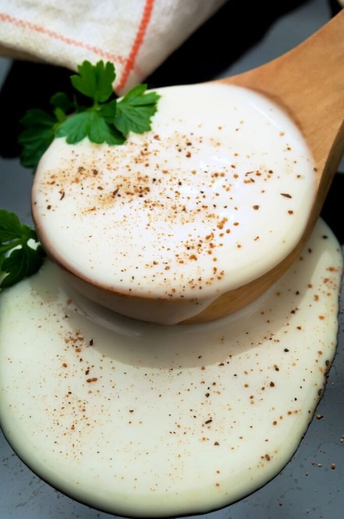 Creamy white sauce with sprinkled spices on a wooden spoon, garnished with fresh parsley leaves.