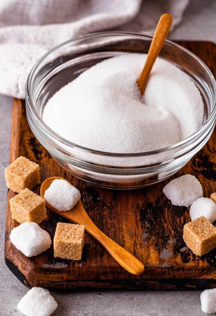 Glass bowl of white sugar with wooden spoon, surrounded by brown and white sugar cubes on a wooden board.