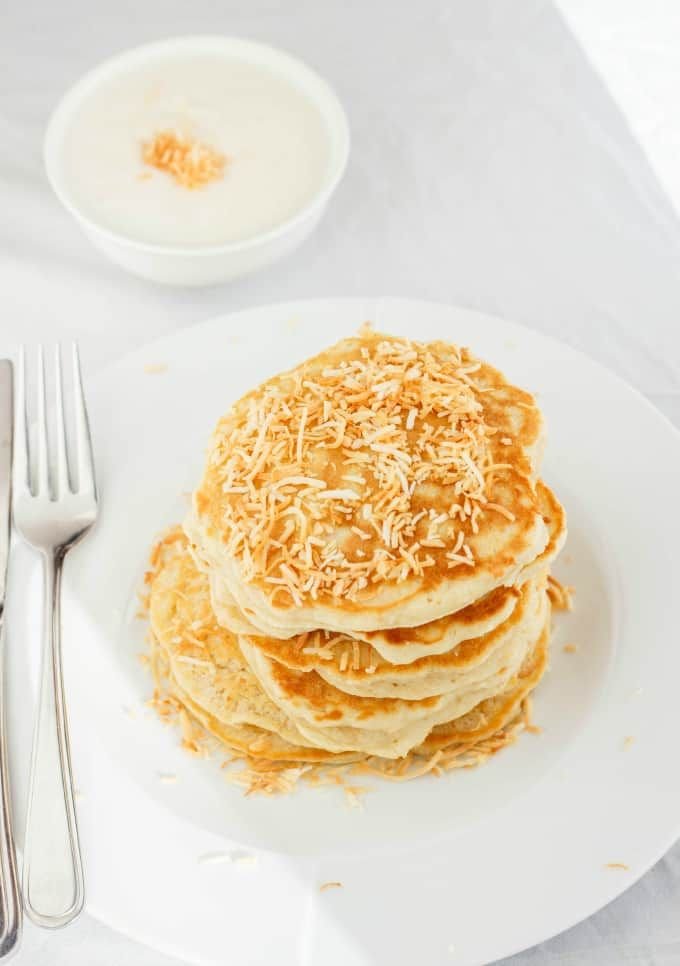 Stack of fluffy pancakes topped with toasted coconut on a white plate, served with a side of creamy coconut sauce.