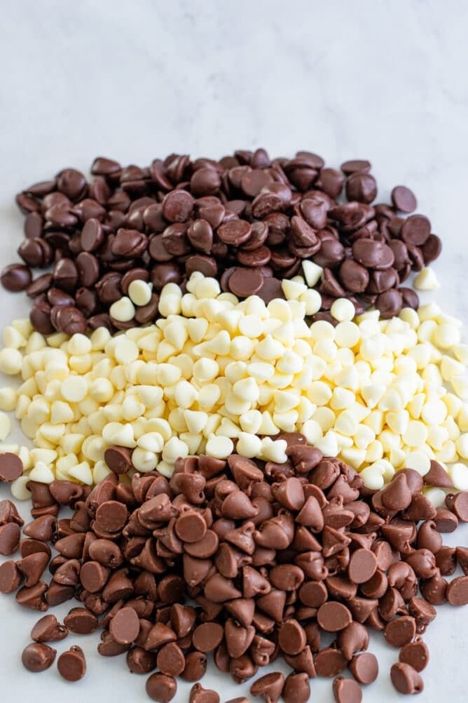 Piles of dark, white, and milk chocolate chips arranged on a marble surface.