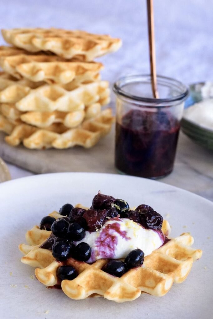 Delicious waffle topped with blueberries and cream, served with a side of berry jam.