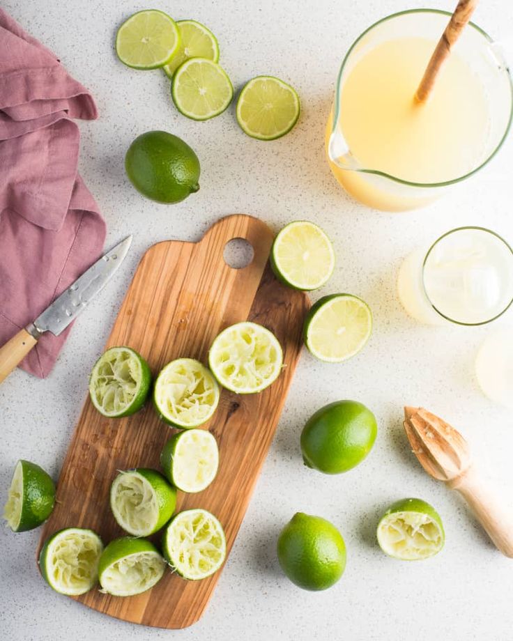 Freshly squeezed lime halves on a cutting board with juice in a pitcher, knife, and wooden reamer on a countertop.