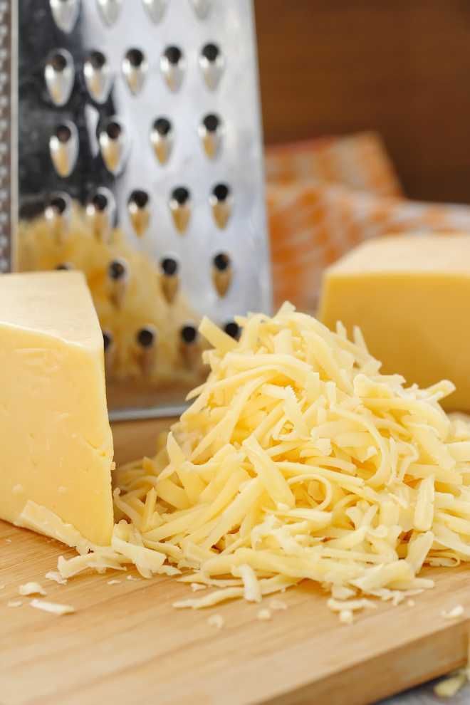 Grated cheese on wooden board with cheese grater in background, perfect for cooking and recipes.