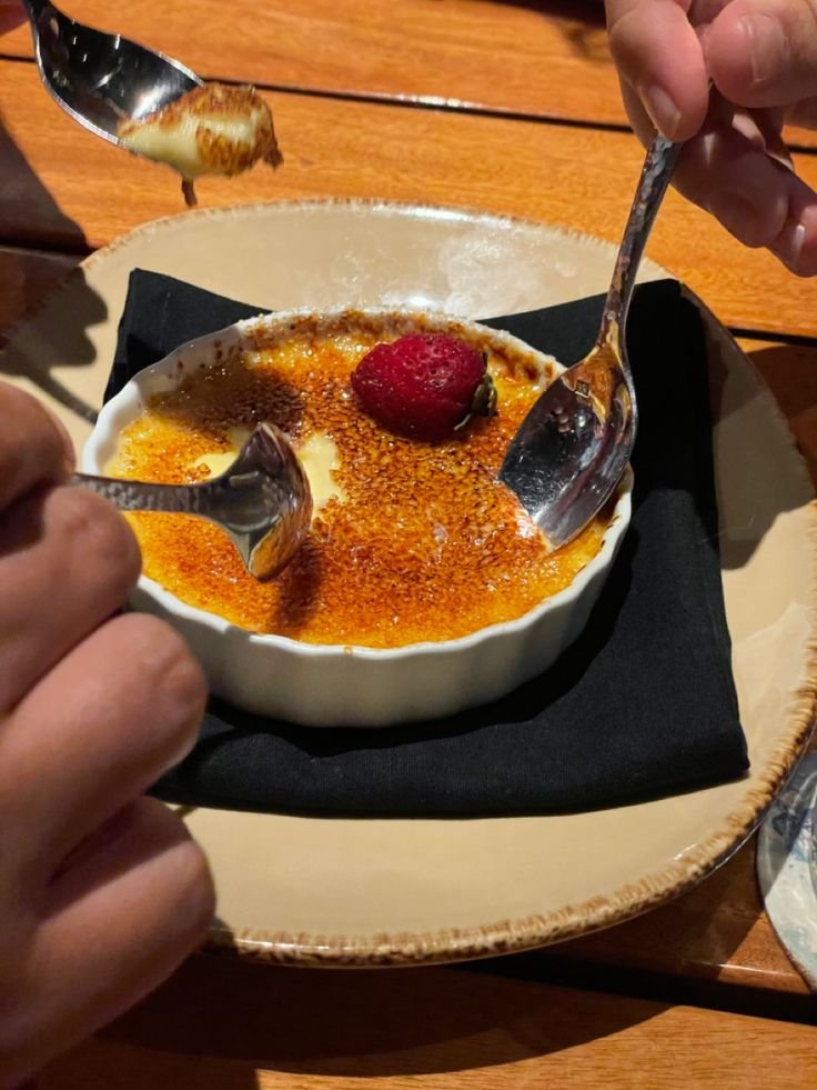 Two spoons digging into a delicious crème brûlée topped with a strawberry on a wooden table.