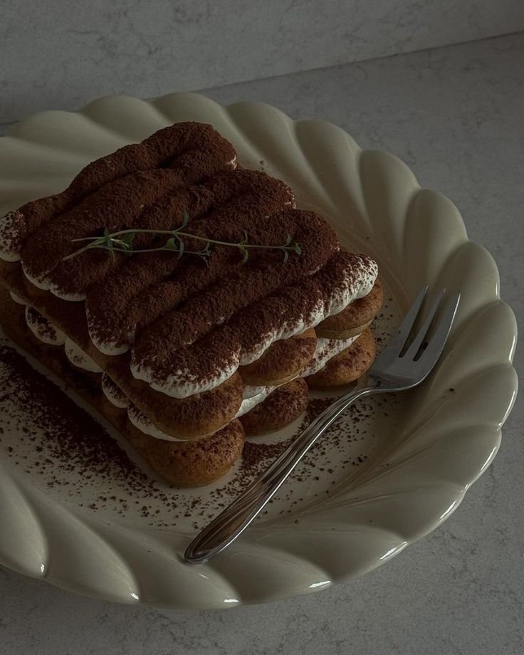 Tiramisu dessert on plate, topped with cocoa powder and garnish, next to a fork on a stone countertop.