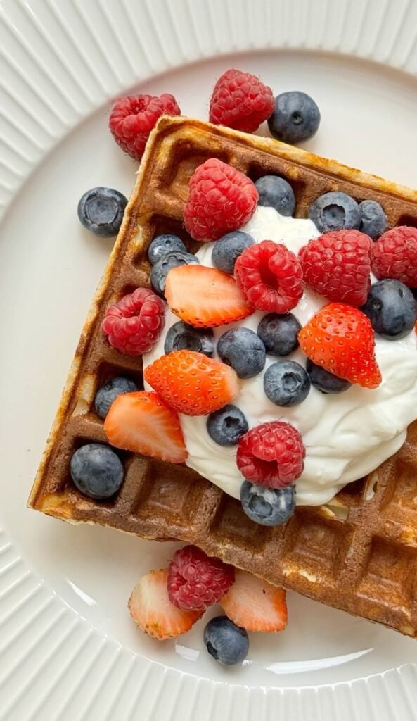 Close-up of waffles with cream, topped with fresh raspberries, strawberries, and blueberries on a white plate.