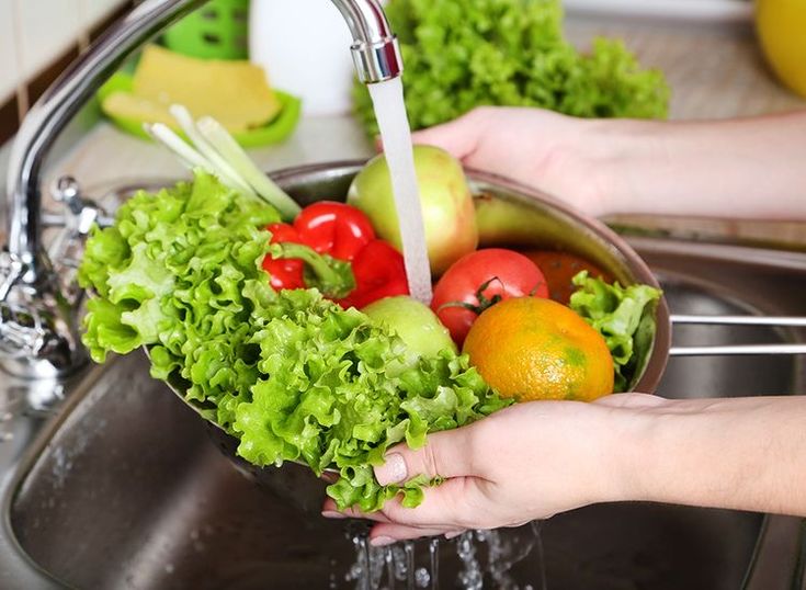 Person rinsing fresh vegetables and fruits under tap water in a kitchen sink.
