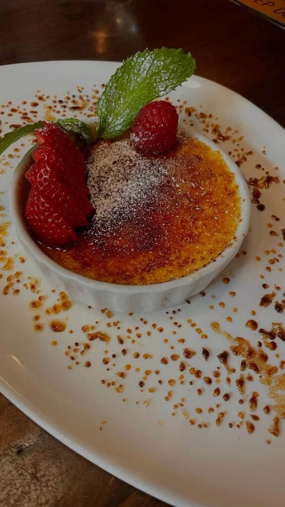 Crème brûlée topped with strawberries, raspberry, mint, and powdered sugar on a white plate.