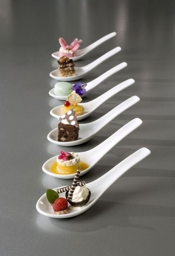 Gourmet desserts arranged on spoons, featuring macaron, cake, and chocolate treats on a sleek gray surface.