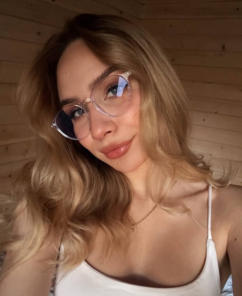 Woman with glasses and wavy blonde hair poses in a wooden room, wearing a white tank top.