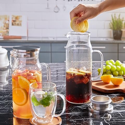 Hand squeezing lemon into pitcher of iced tea on marble counter, surrounded by fresh fruits and a jug of fruit-infused water.