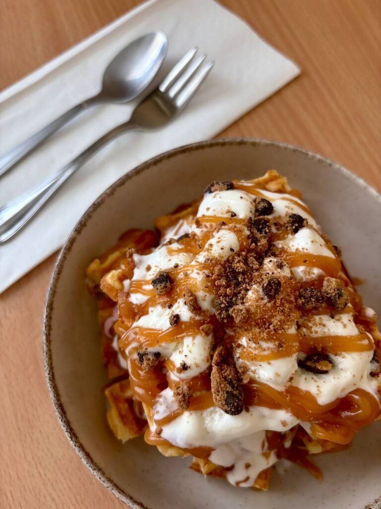 Delicious caramel waffle topped with whipped cream and crumbled cookies on a table with cutlery nearby.