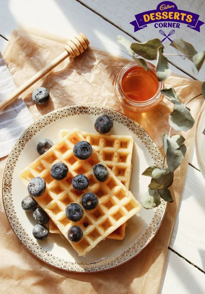 Golden waffles topped with blueberries and honey on a decorative plate under sunlight.