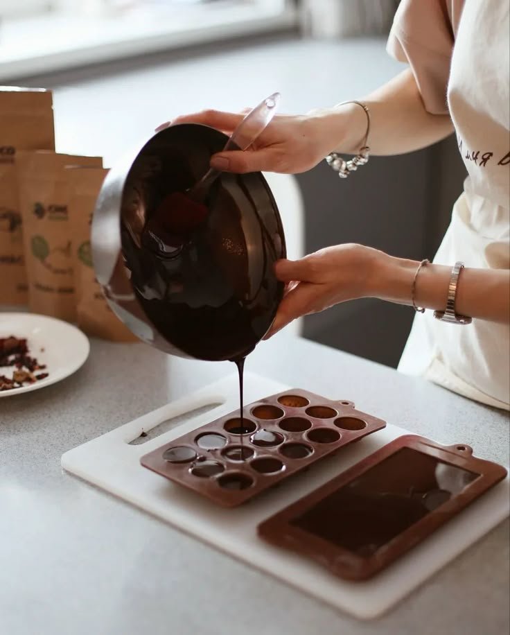 Person pouring liquid chocolate into silicone molds on a kitchen counter.