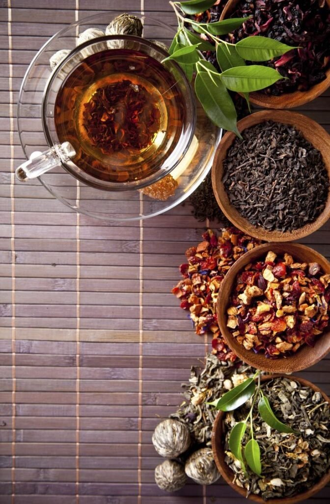 Assorted herbal teas and leaves in wooden bowls with a glass teacup on a bamboo mat background.