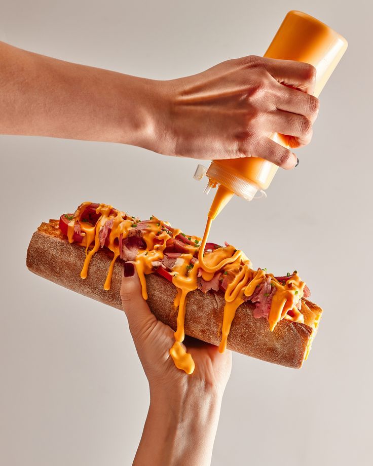 Hand pouring cheese sauce on sub sandwich, topped with meats and herbs. Delicious food preparation close-up.