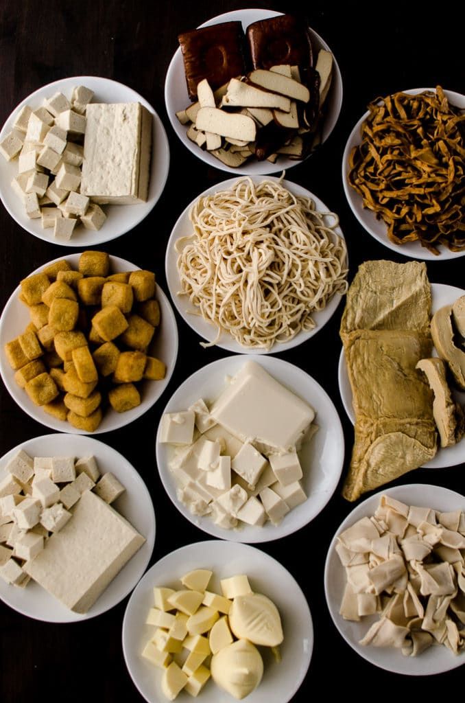 Assorted tofu and mushrooms on plates, including cubes, slices, noodles, and chunks, displayed on a dark background.