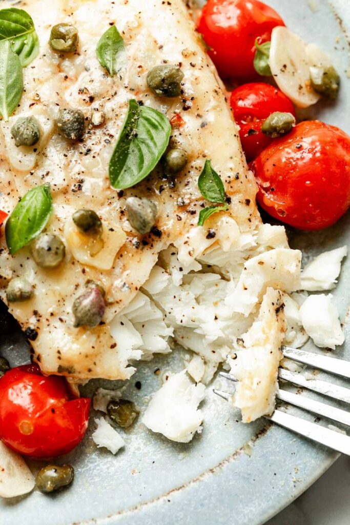Grilled white fish with capers, cherry tomatoes, basil, and garlic on a plate, garnished with ground pepper.
