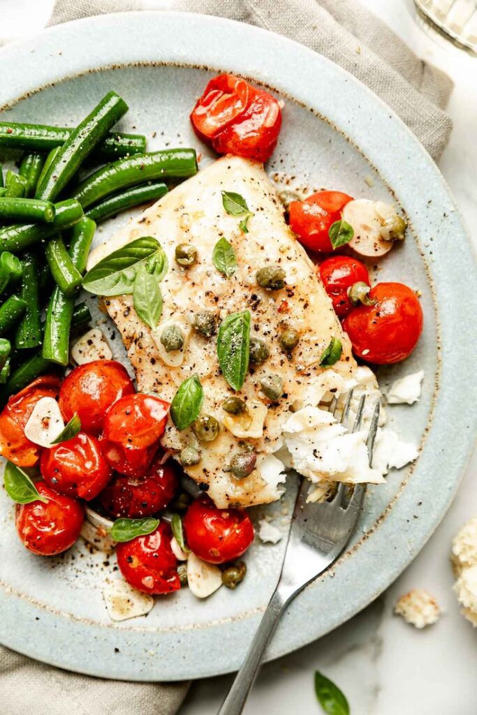 Grilled fish with capers, cherry tomatoes, and green beans on plate, garnished with basil leaves, fork beside.
