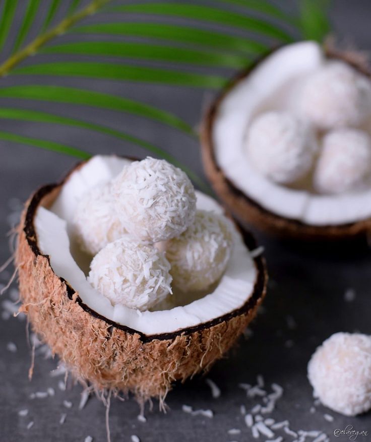 Coconut balls in halved coconut shell with green leaf background, sprinkled with coconut flakes.