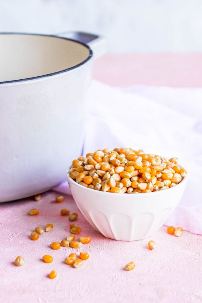 Bowl of yellow popcorn kernels next to a pot on a pink surface, ready for popping.