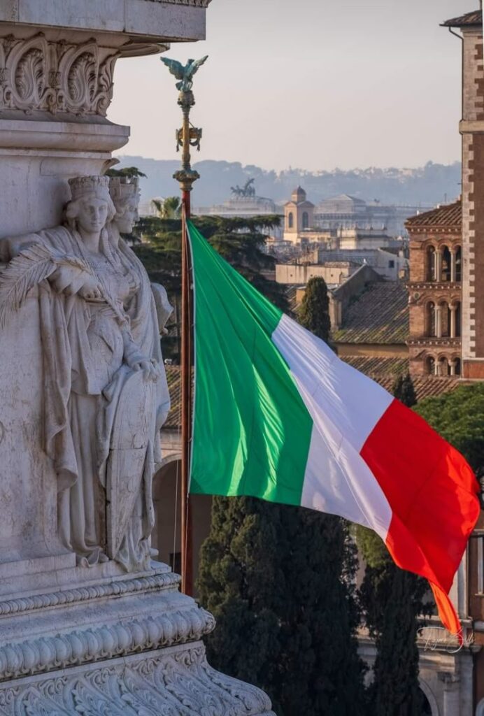 Italian flag waving near a stone statue, with a view of Rome's historic architecture in the background.