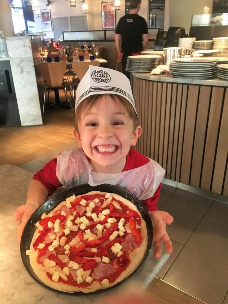Smiling child holding a homemade pizza with cheese and peppers in a cozy restaurant setting.