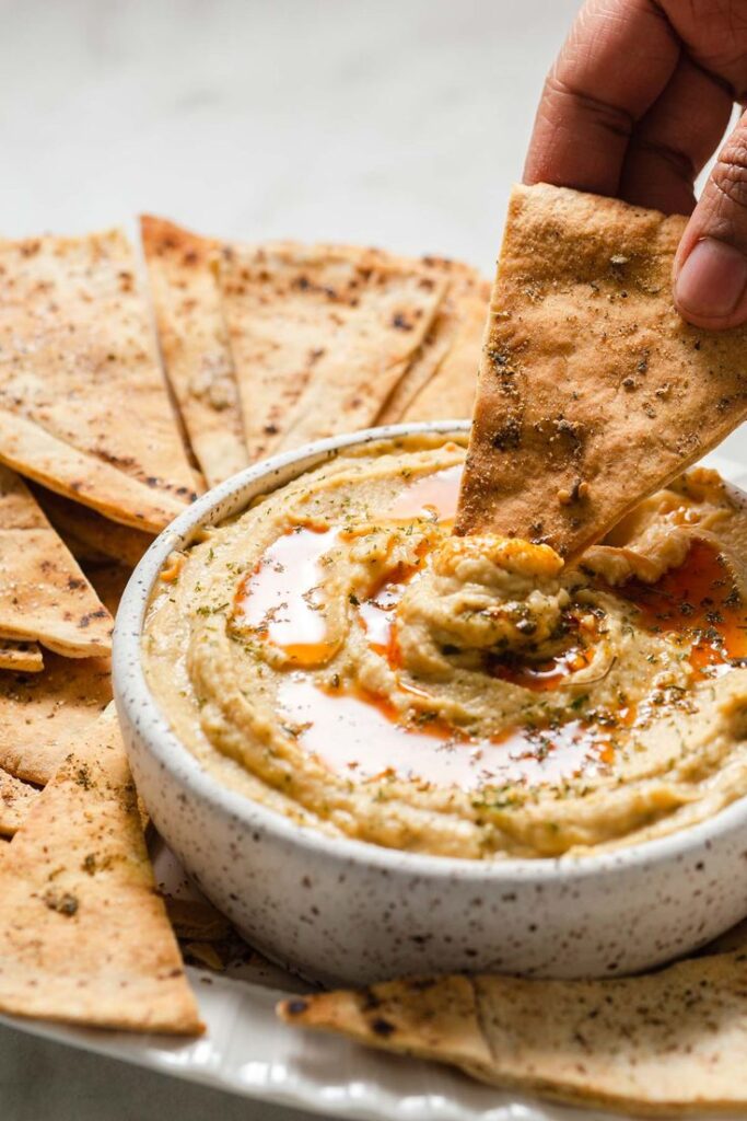 Hand dipping pita chip into a bowl of creamy hummus with paprika and herbs, surrounded by toasted pita slices.