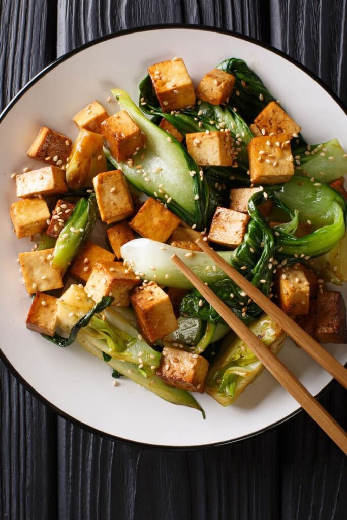 Stir-fried tofu and bok choy with sesame seeds on a white plate with chopsticks, healthy vegan dish.