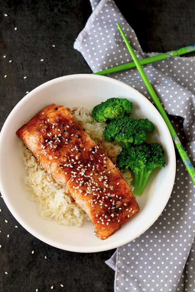 Grilled salmon with sesame seeds on rice, accompanied by broccoli, in a white bowl with chopsticks on a polka dot napkin.