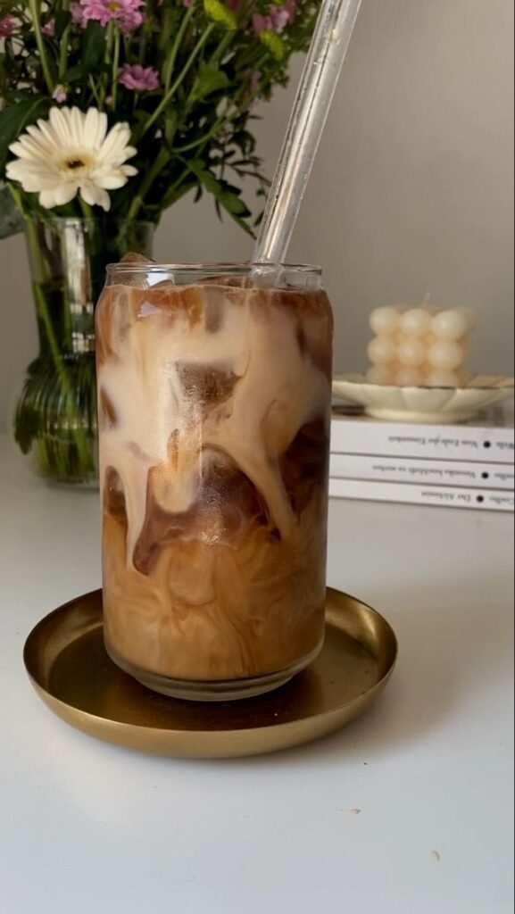 Iced latte with swirling cream in a glass, set on a saucer with flowers and books in the background.