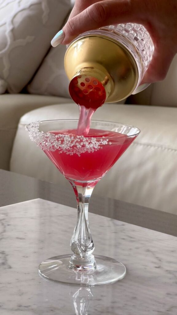 Pouring a vibrant pink cocktail into a sugar-rimmed martini glass on a marble surface.