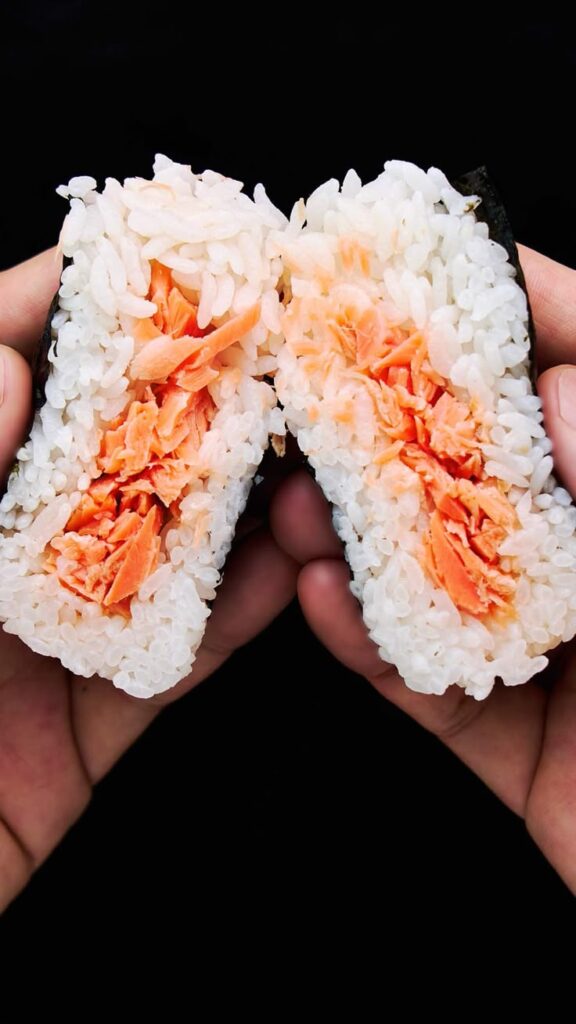 Sliced rice ball with salmon filling held in hands, black background.