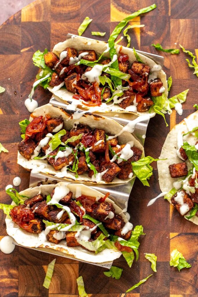 Delicious vegan tacos with grilled tofu, caramelized onions, fresh lettuce, and creamy sauce on a wooden board.