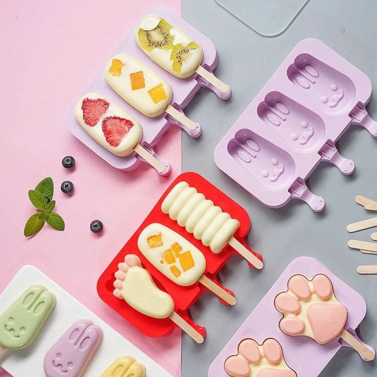 Colorful homemade popsicles with fruit in cute molds, surrounded by ingredients and sticks on a pastel surface.