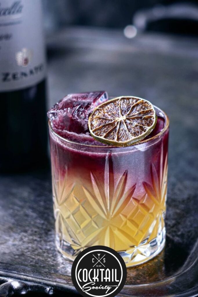 Elegant cocktail with layered colors and dried citrus garnish in a textured glass, alongside a wine bottle.