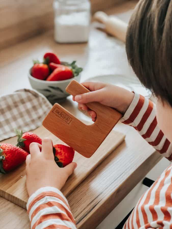 Child using a wooden knife to cut strawberries on a kitchen countertop.