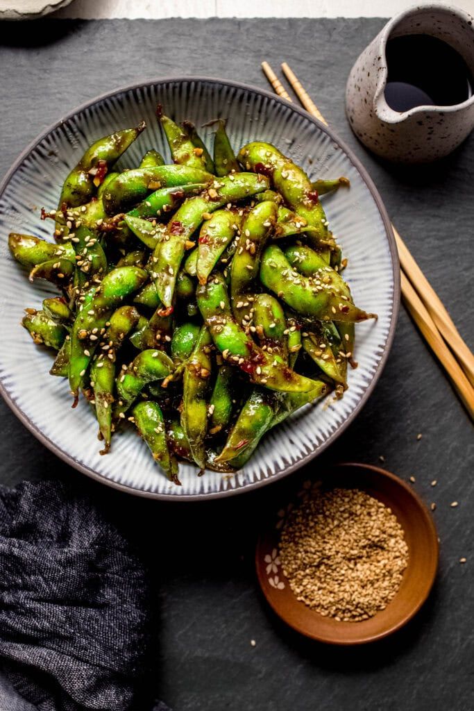Spicy garlic edamame with sesame seeds served in a bowl with soy sauce and chopsticks on a slate table.