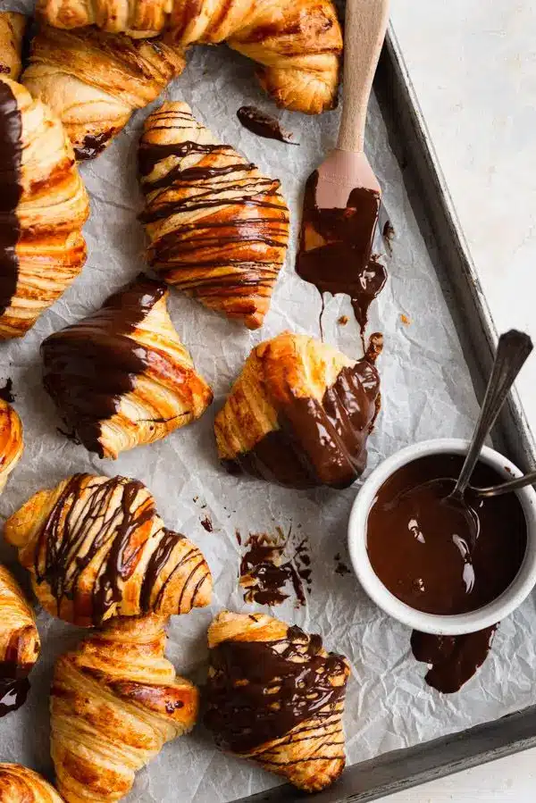 Fresh croissants drizzled with rich chocolate on a baking tray, beside a bowl of melted chocolate.