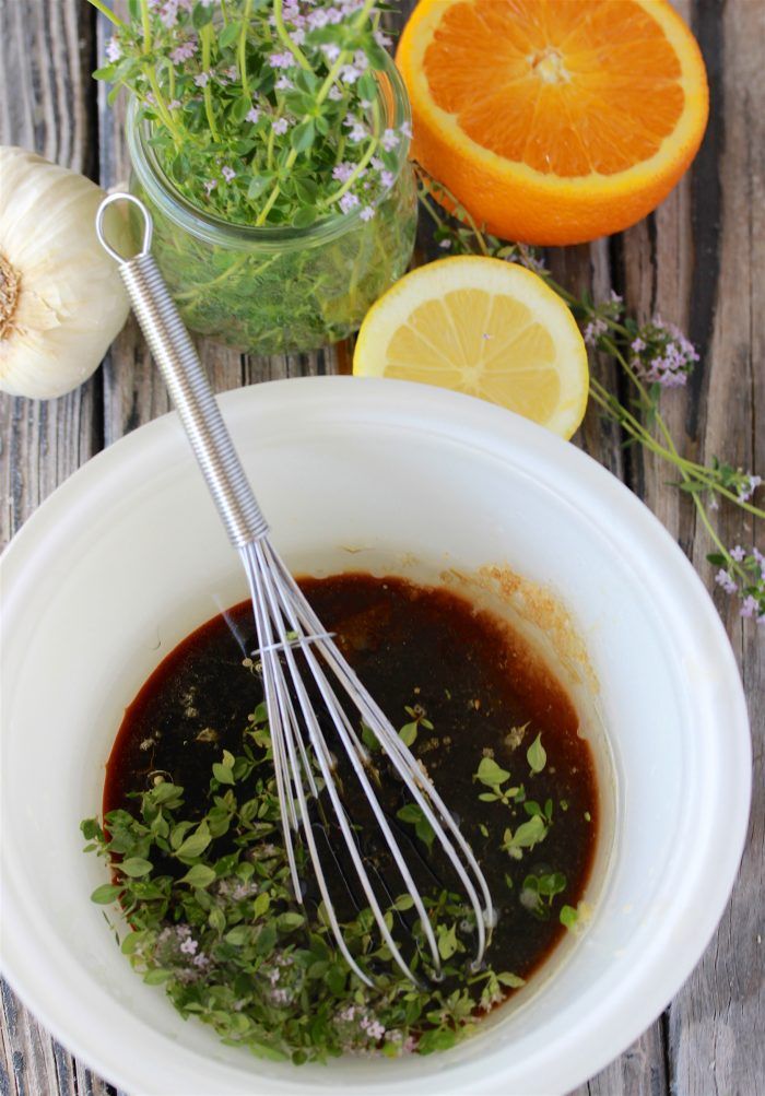 Whisk in a bowl of marinade with fresh herbs, garlic, orange, and lemon on a rustic wooden table.