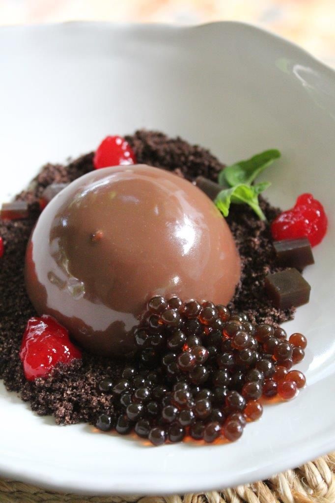 Chocolate mousse dessert with berry pearls and mint garnish on a plate, accented with chocolate crumbs and fruit jelly.